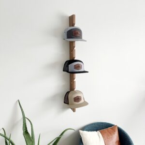 3 hats displayed on a wood hat rack hung on a white wall with plant and chair in picture